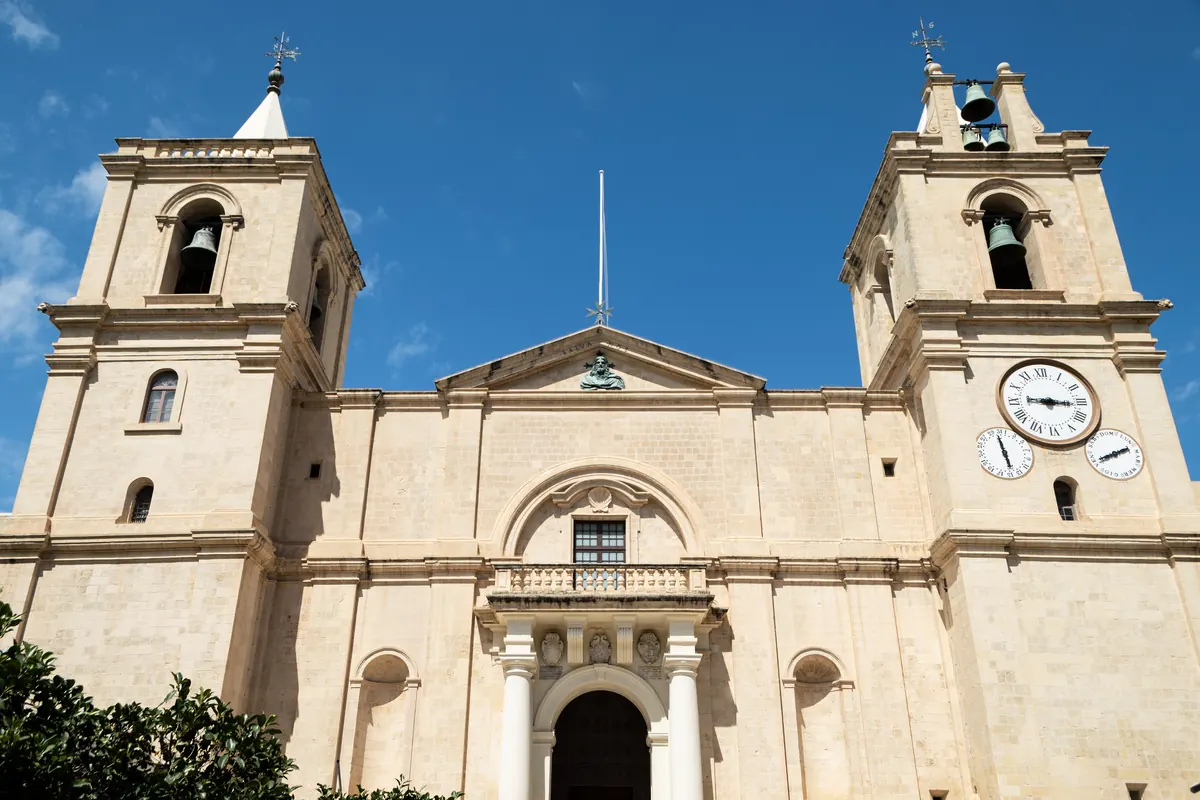 The plain limestone front of St John's Co-Cathedral in Valletta, with two bell towers, a balcony over the doorway and a blue sky behind